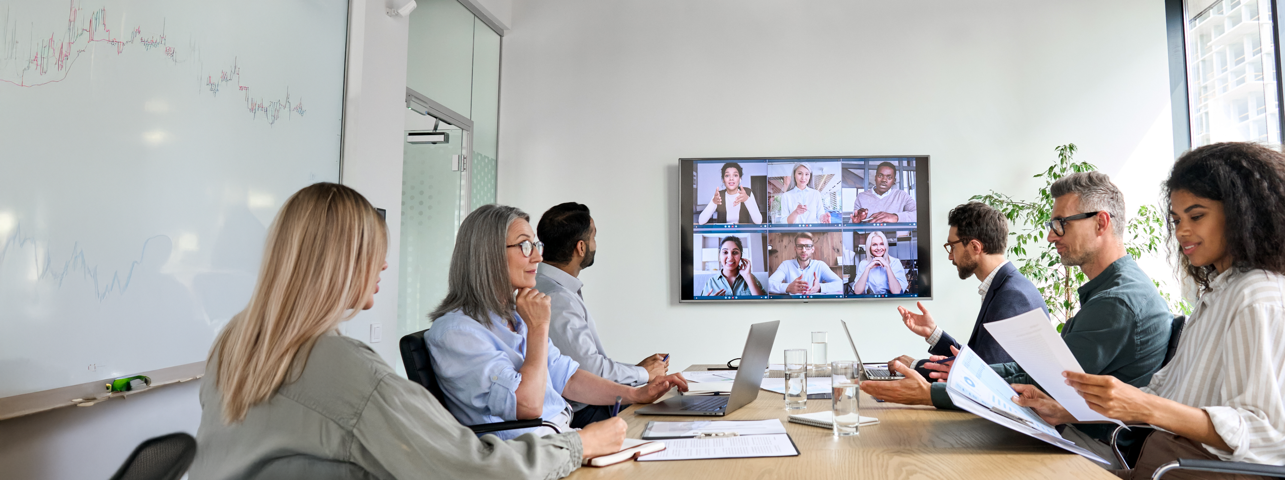 Four people sit speaking to each other in a conference room at a table with water glasses and an open laptop. A screen on the wall shows six other people participating in a video call. A text box next to the image reads, “Fios Business Internet. Connection at the speed of light.”